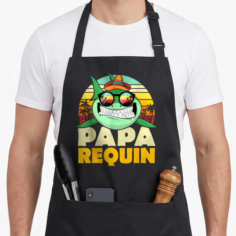 Tablier de cuisine noir avec un requin vert portant un sombrero et des lunettes reflétant des palmiers.