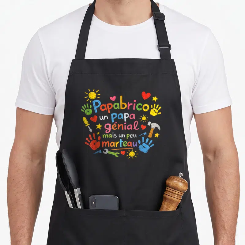 Tablier de cuisine noir avec inscription Papabrico un papa génial mais un peu marteau entouré de mains et d'outils colorés.