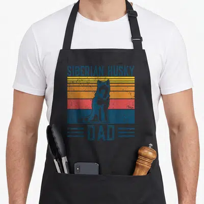 Tablier de cuisine noir avec illustration rétro d'un husky sibérien assis sur des bandes colorées orange et bleues.