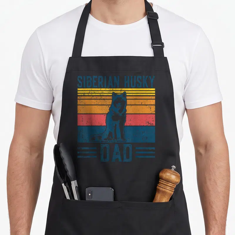 Tablier de cuisine noir avec illustration rétro d'un husky sibérien assis sur des bandes colorées orange et bleues.