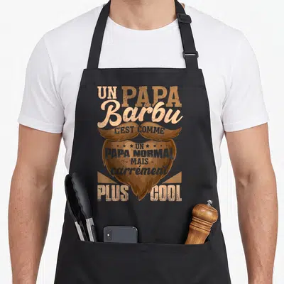 Tablier de cuisine noir avec une illustration de barbe et moustache et le texte un papa barbu c'est comme un papa normal mais carrément plus cool