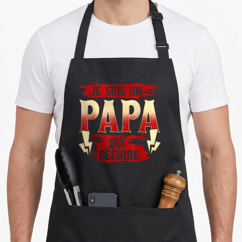 Tablier de cuisine noir avec inscription Je suis un papa qui déchire en lettres rouges et jaunes stylisées.