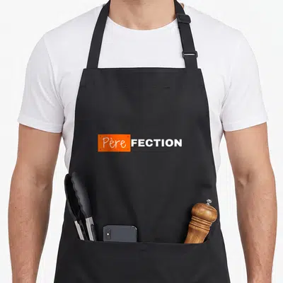 Tablier de cuisine noir avec une étiquette orange portant l'inscription Père-fection en lettres blanches.