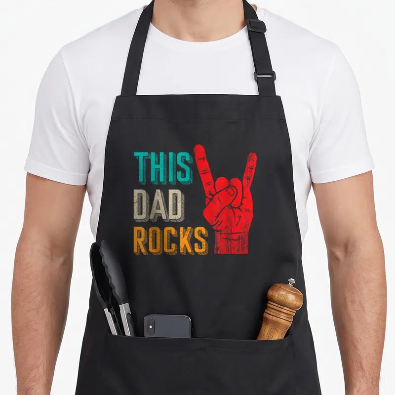 Tablier de cuisine noir avec inscription This Dad Rocks et main faisant le signe du rock en rouge.