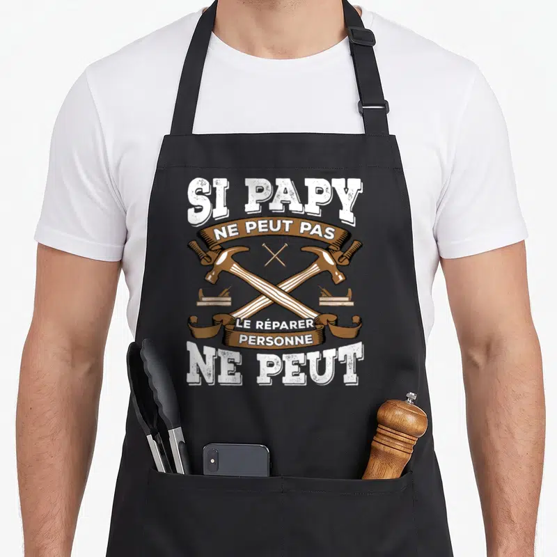 Tablier de cuisine noir avec deux marteaux croisés et l'inscription Si papy ne peut pas le réparer personne ne peut en blanc et marron.