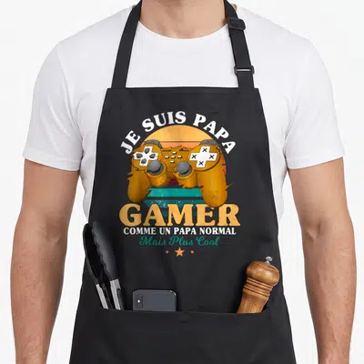 Tablier de cuisine noir avec une manette de jeu vidéo orange et le texte Je suis papa gamer comme un papa normal mais plus cool.