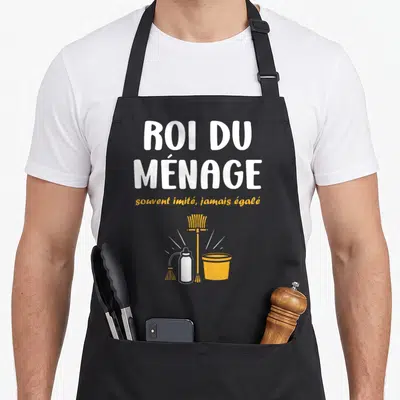 Tablier de cuisine noir avec texte Roi du ménage souvent imité jamais égalé et ustensiles de nettoyage jaune et blanc.