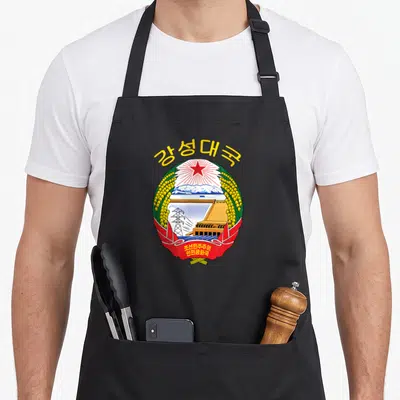 Tablier de cuisine noir avec un emblème circulaire coloré représentant un barrage, une étoile rouge et des épis de riz dorés.