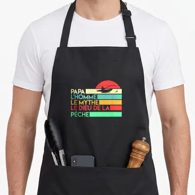 Tablier de cuisine noir avec motif vintage couleur affichant Papa l'homme le mythe dieu de la pêche sur fond de soleil rouge.