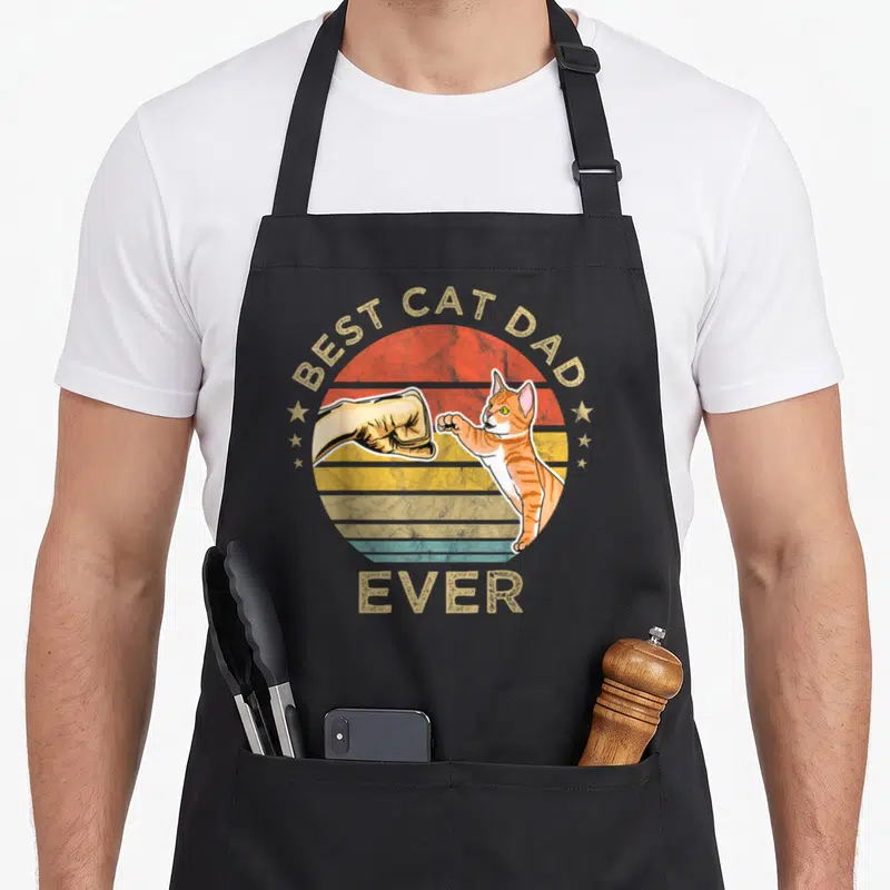 Tablier de cuisine noir avec illustration d'un chat orange et d'une main en poing sur fond circulaire coloré.