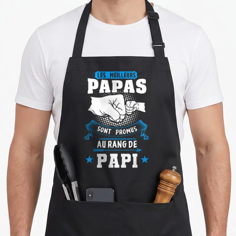 Tablier de cuisine noir avec poings serrés blancs et texte Les meilleurs Papas sont promus au rang de Papi en blanc et bleu.