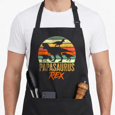 Tablier de cuisine noir avec silhouette de dinosaure et texte Papasaurus Rex sur fond de soleil coloré vintage.
