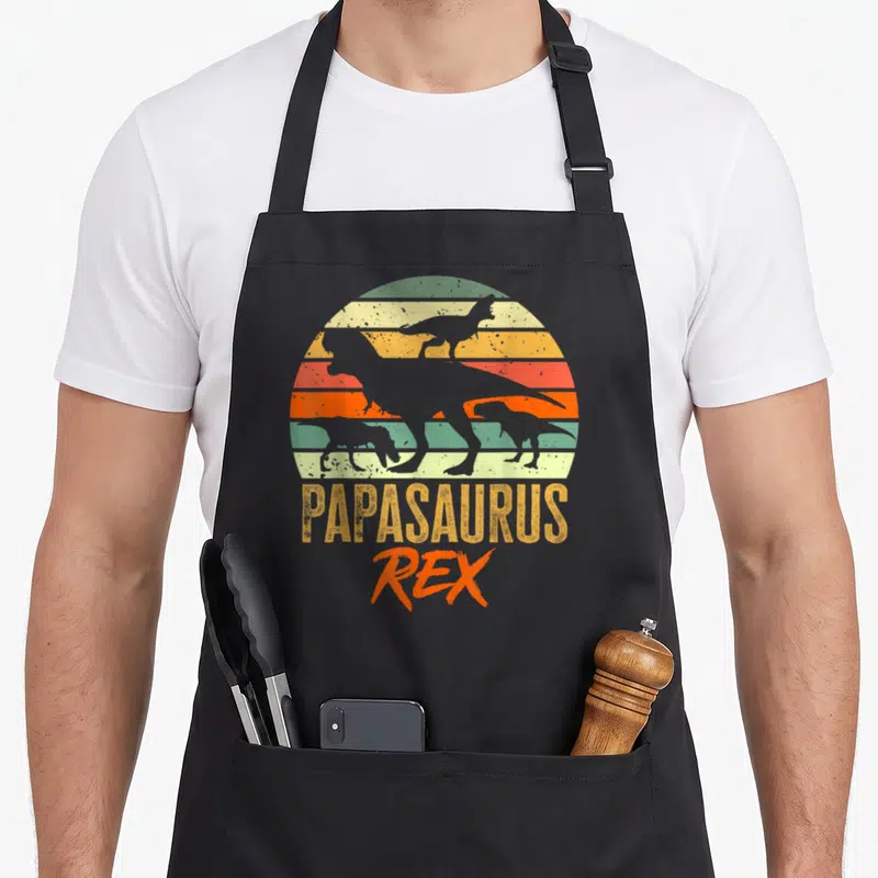 Tablier de cuisine noir avec silhouette de dinosaure et texte Papasaurus Rex sur fond de soleil coloré vintage.