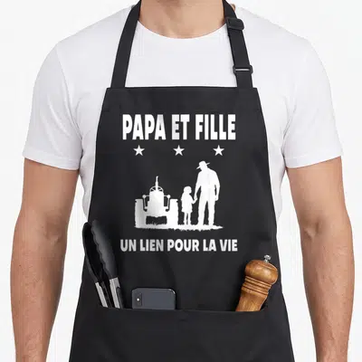 Tablier de cuisine noir avec silhouette blanche d'un homme tenant la main d'une enfant près d'un tracteur et du texte Papa et Fille.