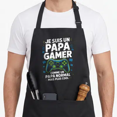 Tablier de cuisine noir avec une illustration de manette de jeu vidéo bleue et verte entourée de texte blanc humoristique.