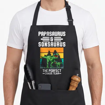 Tablier de cuisine noir avec deux dinosaures verts aux lunettes de soleil et le texte Papasaurus et Sonsaurus équipe chaos parfait.