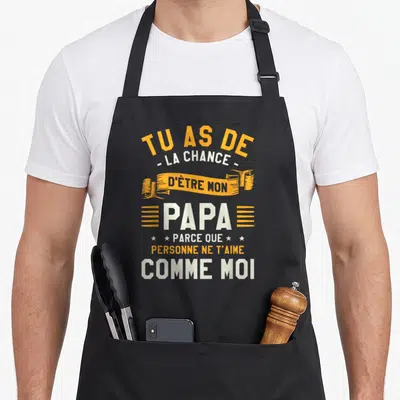Tablier de cuisine noir avec inscription humoristique sur le thème familial et paternel en lettres orange et blanches.
