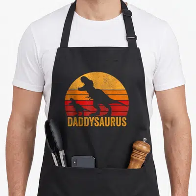 Tablier de cuisine noir avec silhouette de dinosaures et texte Daddysaurus sur fond de soleil coloré.