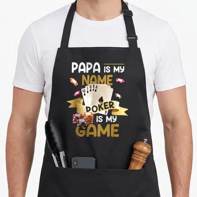 Tablier de cuisine noir avec une illustration de cartes à jouer et des jetons de poker sur fond gris uni.