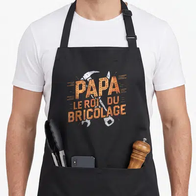 Tablier de cuisine noir avec texte Papa le roi du bricolage et outils croisés en orange et blanc.