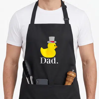 Tablier de cuisine noir avec un canard jaune portant un chapeau haut de forme gris et le mot Dad imprimé en blanc.