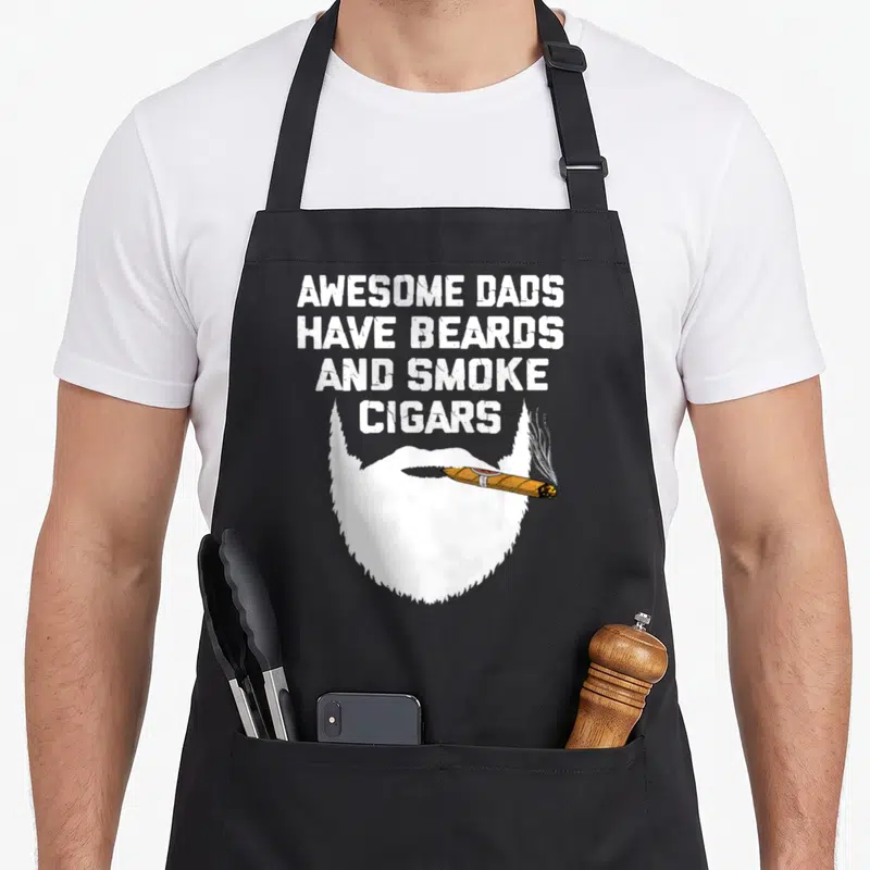 Tablier de cuisine noir avec une barbe blanche stylisée tenant un cigare et du texte en anglais blanc au-dessus.