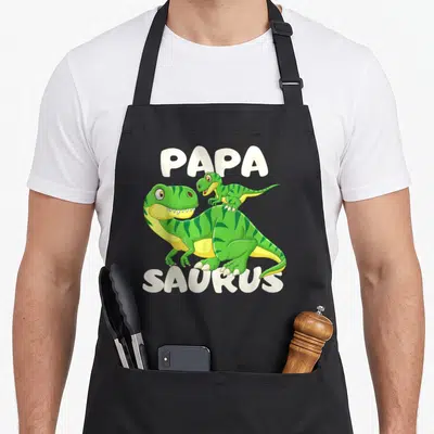 Tablier de cuisine noir avec un dinosaure vert et son petit, les mots Papa Saurus en blanc.