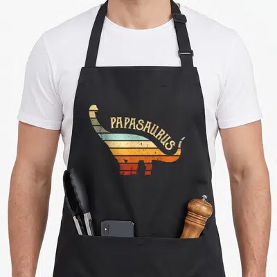 Tablier de cuisine noir avec silhouette de dinosaure multicolore et texte PAPASAURUS en lettres capitales vieillies.