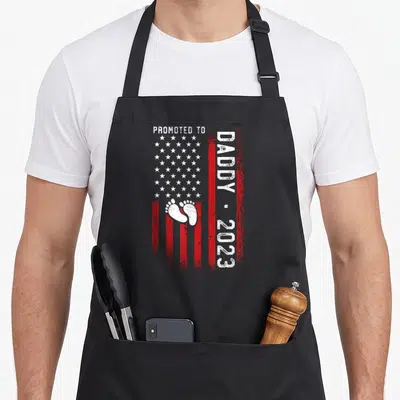 Tablier de cuisine noir avec motif « Promoted to Daddy 2023 » étoiles et empreintes de pieds.