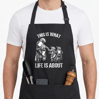 Tablier de cuisine noir avec illustration blanche d'un père motocross tendant la main à deux enfants sur mini-motos.