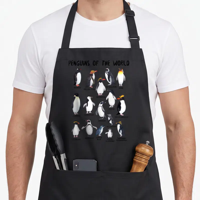 Tablier de cuisine noir avec une illustration colorée présentant divers manchots comme l'empereur, le macaroni et le gentoo.