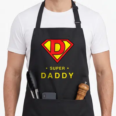 Tablier de cuisine noir avec emblème rouge jaune D et texte SUPER DADDY en lettres capitales jaunes.