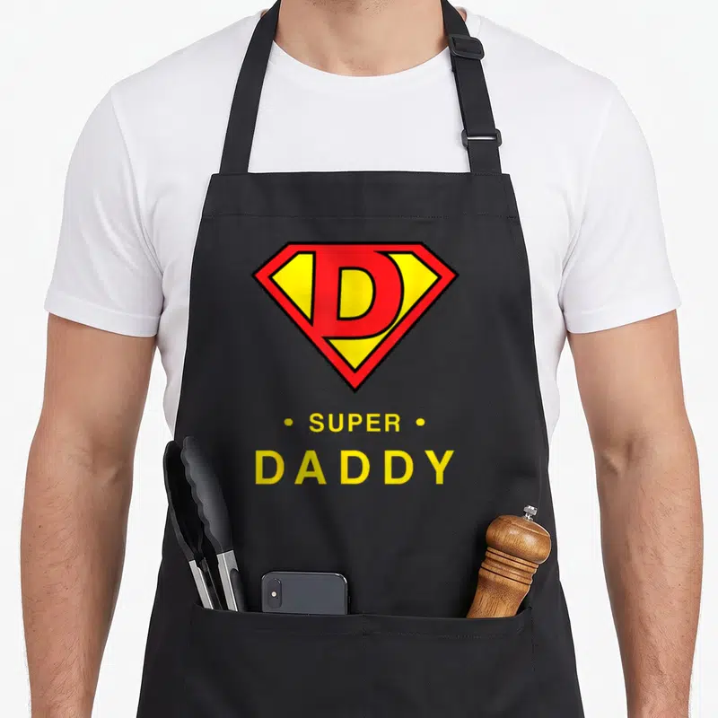 Tablier de cuisine noir avec emblème rouge jaune D et texte SUPER DADDY en lettres capitales jaunes.