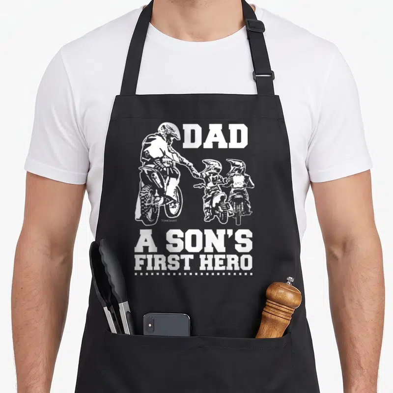 Tablier de cuisine noir avec une illustration blanche d'un père et deux enfants en tenue de motocross, le texte DAD A SON'S FIRST HERO en gros caractères.