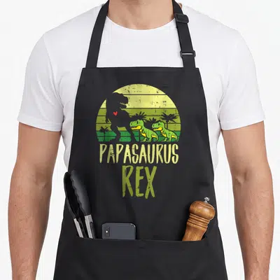Tablier de cuisine noir avec illustration vintage Papasaurus Rex et deux petits dinosaures verts devant un soleil jaune.