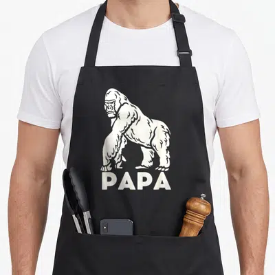 Tablier de cuisine noir avec illustration blanche d'un gorille debout au-dessus du mot PAPA en lettres capitales.