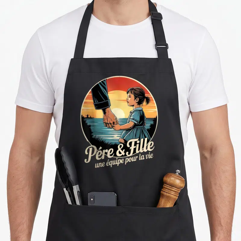 Tablier de cuisine noir avec illustration circulaire d'une main d'adulte tenant celle d'une petite fille devant un coucher de soleil sur l'eau.