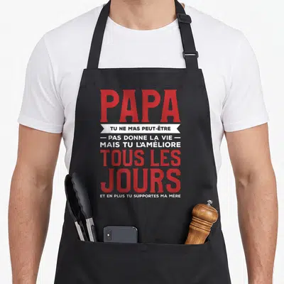 Tablier de cuisine noir avec typographie rouge et blanche sur fond gris, message humoristique dédié au papa.