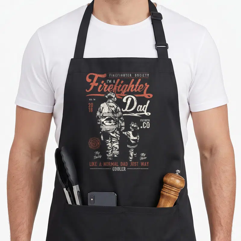Tablier de cuisine noir avec silhouette de pompier et enfant en tenue de feu, texte Firefighter Dad en rouge et blanc.
