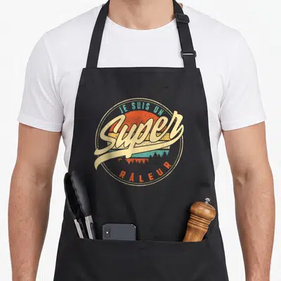 Tablier de cuisine noir avec logo circulaire retro affichant Je suis un super râleur sur fond de soleil orange et turquoise.