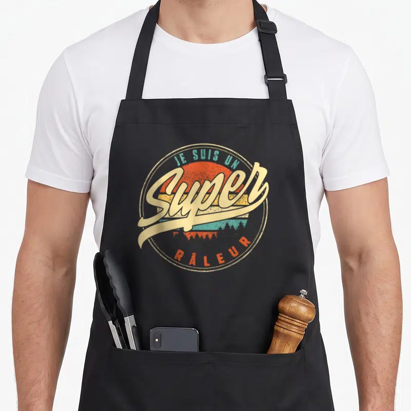 Tablier de cuisine noir avec logo circulaire retro affichant Je suis un super râleur sur fond de soleil orange et turquoise.