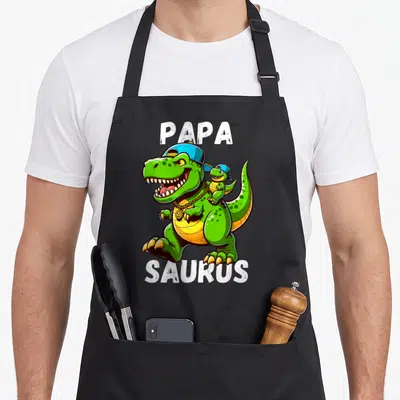 Tablier de cuisine noir avec un T Rex vert portant une casquette bleue et le texte Papa Saurus imprimé en blanc.