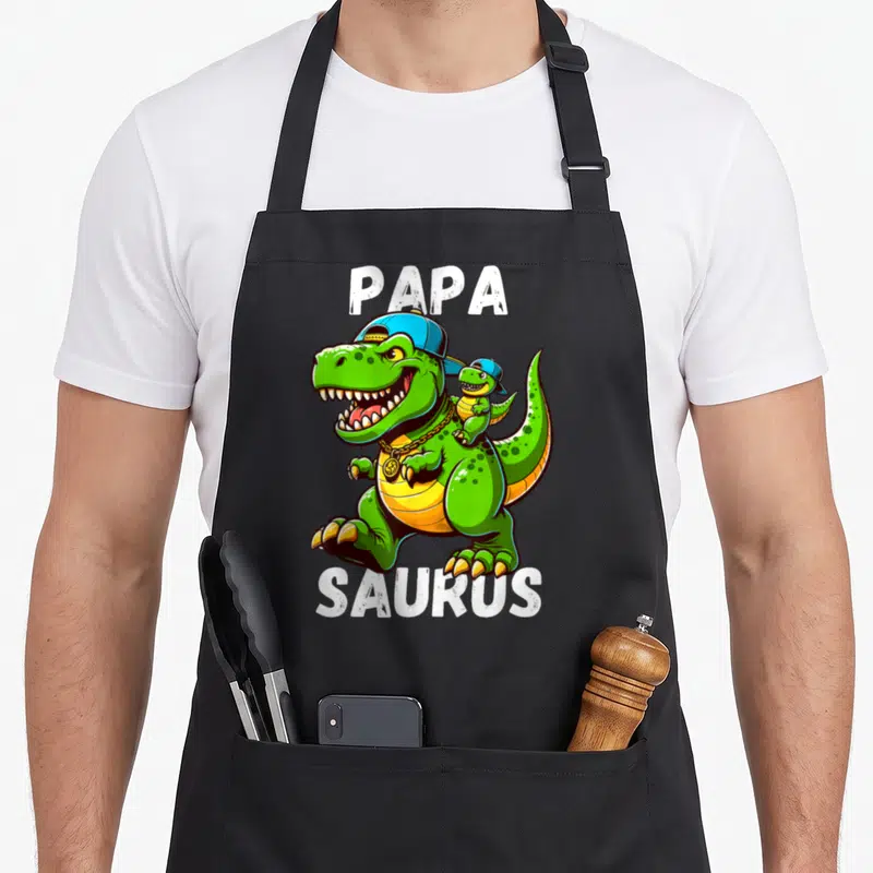 Tablier de cuisine noir avec un T Rex vert portant une casquette bleue et le texte Papa Saurus imprimé en blanc.