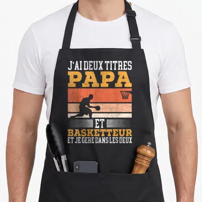 Tablier de cuisine noir avec silhouette basketteur et texte J'ai deux titres Papa et basketteur en orange et blanc.