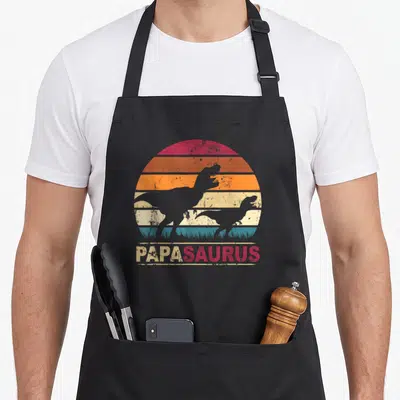 Tablier de cuisine noir avec illustration vintage Papa Saurus Rex et silhouettes de dinosaures sur fond coloré.