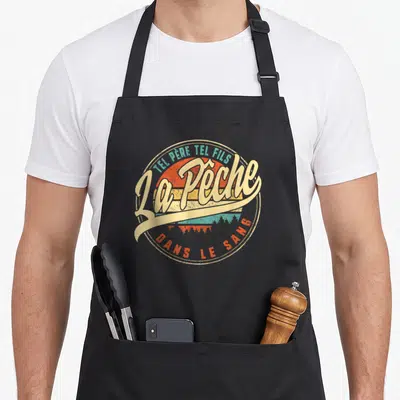 Tablier de cuisine noir avec un logo circulaire vintage affichant La Pêche Dans Le Sang sur fond de soleil couchant.