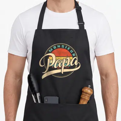 Tablier de cuisine noir avec logo rond Monsieur Papa style rétro soleil couchant multicolore et texte calligraphié beige.
