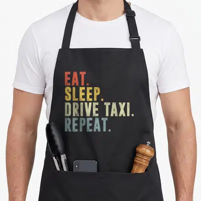 Tablier de cuisine noir avec inscription EAT SLEEP DRIVE TAXI REPEAT en lettres orange jaune beige et bleu sur fond uni.