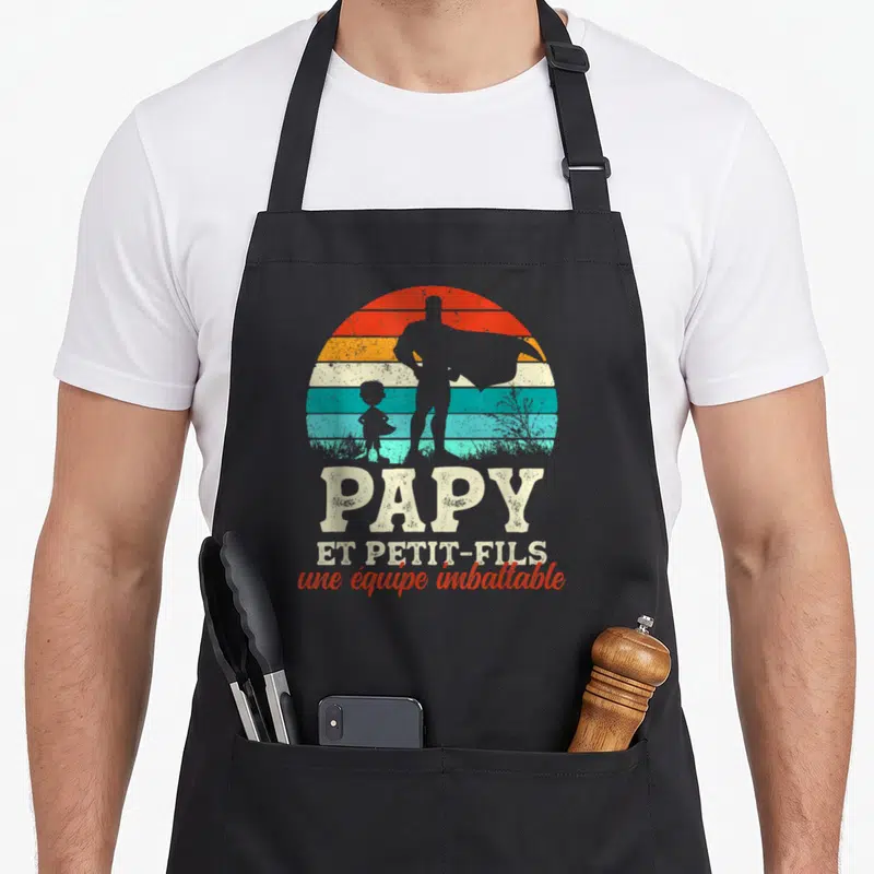 Tablier de cuisine noir avec silhouette super-héros et enfant devant un soleil rayé multicolore.