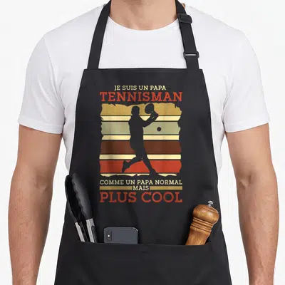 Tablier de cuisine noir avec silhouette de joueur de tennis et texte Je suis un papa tennisman plus cool en lettres orange et beige.